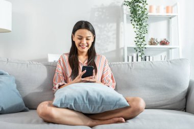 Kanepede oturan gülümseyen kadın yastıklı akıllı telefon kullanıyor.