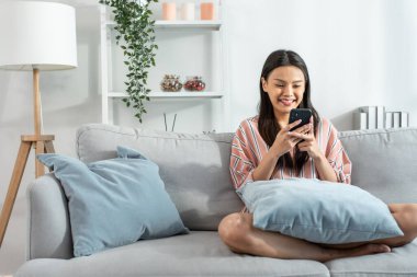 Asyalı güzel kadın kanepede oturur ve cep telefonuyla sohbet eder. Mutlu çekici genç kız evde vakit geçirir, rahatlar, iletişim kurar ve oturma odasında akıllı telefondan tartışır..