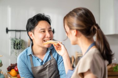 Asyalı genç bir kadın mutfakta ekmek tutuyor ve erkeğe veriyor. Çekici erkek ve kadın çift önlük giyer, evde sağlık hizmetleri için kahvaltı hazırlar. Diyet ve Sağlıklı Yemek Konsepti