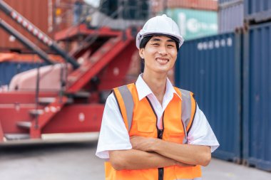 Genç Asyalı mühendisin portresi konteynır limanında çalışıyor. İthalat ihracat için kargo gemisindeki depo lojistiğinde planlamaları için çekici erkek işçiler ve ürünler..