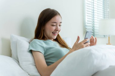 Beyaz bir kadın sabahları cep telefonu sohbetini evde yapıyor. Mutlu, sıradan, güzel bir kadın yatak odasında uzanıyor. Tatil haftasonunun tadını çıkarıyor. Akıllı telefon kullanarak evdeki teknolojiyle iletişim kuruyor..