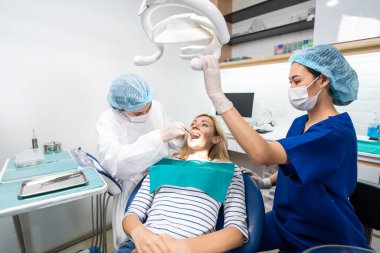 Kadın diş hekimi, diş sağlığı kliniğindeki beyaz dişli kızı muayene ediyor. Diş hekimliği koltuğunda yatan çekici kadın hastanede muayene sırasında doktordan diş tedavisi görüyor.