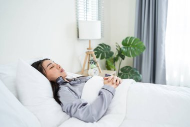Asyalı genç ve güzel bir kız evdeki yatak odasında mutlulukla uyuyor. Pijamalı çekici bir kadın. Evde sabahları rahat battaniye ve yastıkla yatarken mutlu ve rahatlamış hissediyor..