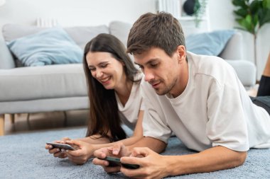 Beyaz tenli çekici çift oturma odasında akıllı telefon oyunu oynuyor. Çekici erkek ve kadın mutlu ve rahatlamış hissediyorlar, boş zamanlarını evde birlikte oyun oynayarak geçiriyorlar..