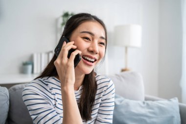 Asyalı güzel kadın kanepede oturur ve cep telefonuyla konuşur. Mutlu çekici genç kız evde vakit geçirir, rahatlar, iletişim kurar ve oturma odasında akıllı telefondan tartışır..