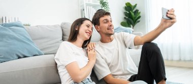 Beyaz tenli çekici çift oturma odasında cep telefonuyla konuşuyor. Çekici erkek ve kadın kendilerini mutlu ve rahat hissediyorlar. Sabahları akıllı telefondan sanal konuşmanın tadını çıkarıyorlar..