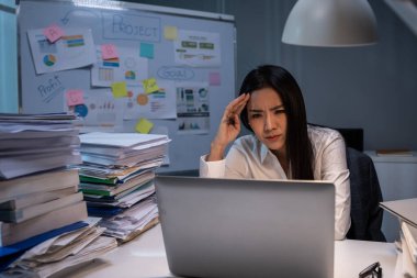 Asyalı sinirli iş kadını geceleri ofiste dizüstü bilgisayar kullanıyor. Çekici profesyonel kadın çalışan masada oturuyor, baş ağrısından midesi bulanıyor ve şirkette bir sürü iş arıyor..