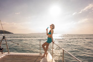 Yatta parti yaparken bikinili bir kadın şampanya içiyor. Birlikte takılan çekici güzel bayan turist yaz günbatımında katamaran teknesiyle açılırken tatil tatilini kutluyor..