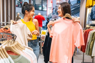 Asyalı iki güzel kadın alışveriş merkezinde kıyafet ürünlerine bakıyor. Çekici, mutlu kız alışveriş merkezinde elbise ve giysi raflarından yeni tişört seçmek için mağazaya girmekten hoşlanıyor.