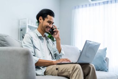 Bilgisayarda yazı yazan ve cep telefonuyla konuşan Asyalı genç işadamı. Çekici serbest çalışan erkek işçi kanepede oturuyor, dizüstü bilgisayarını kullanıyor, evdeki oturma odasında mutlulukla çalışıyor..