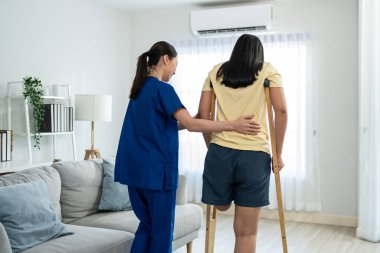 Asyalı kadın bacak ampütesi hemşirenin desteğiyle fizyoterapi yapıyor. Çekici genç kız bacaksız hasta oturma odasında kadın bakıcı doktorla yavaşça yürüme alıştırması yapıyor..