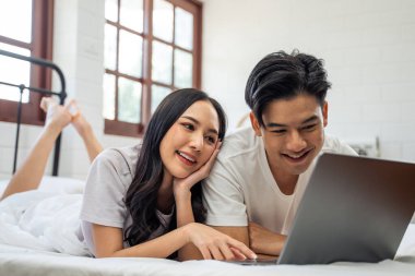 Asyalı çekici kadın sabahları kocasıyla yatakta dizüstü bilgisayar kullanıyor. Yeni evli çiftimiz yatak odasında uyandıktan sonra bilgisayardan film izlerken mutlu ve rahatlamış hissediyorlar.