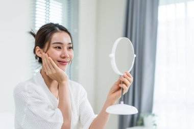 Asyalı güzel kadın yüzüne losyon sürdükten sonra aynaya bakıyor. Yatak odasında yatakta oturan çekici genç kız, sağlıklı güzellik tedavisi ve cilt bakımı için mutlu ve gururlu.