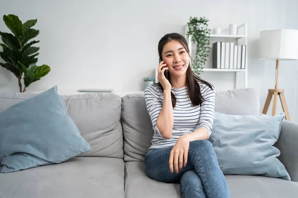 Asyalı güzel kadın kanepede oturur ve cep telefonuyla konuşur. Mutlu çekici genç kız evde vakit geçirir, rahatlar, iletişim kurar ve oturma odasında akıllı telefondan tartışır..