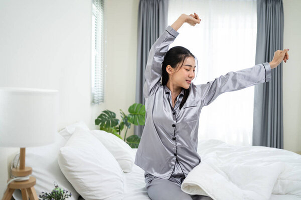 Азиатская красивая девушка в пижаме просыпается утром со счастьем. Привлекательная молодая женщина улыбается, чувствовать себя счастливым и расслабиться, а затем растяжения тела после вставания от сна на кровати в спальне на дому.