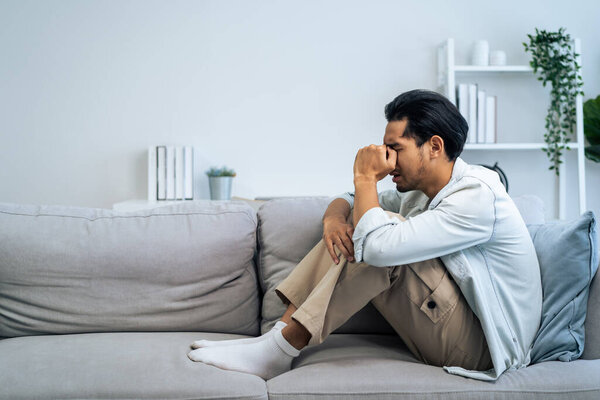 Молодой азиат расстроен тем, что мужчина сидит один дома в гостиной. Привлекательный несчастный мужчина чувствует себя очень грустным, одиноким и разочарованным из-за жизненных проблем. Подумайте о деньгах, потере бюджета, банкротстве..