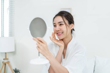 Asyalı güzel kadın yüzüne losyon sürdükten sonra aynaya bakıyor. Yatak odasında yatakta oturan çekici genç kız, sağlıklı güzellik tedavisi ve cilt bakımı için mutlu ve gururlu.