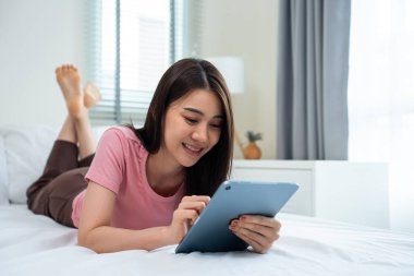 Asyalı çekici kadın sabahları evde yatakta tablet sohbeti yapıyor. Mutlu, sıradan, güzel bir kadın yatak odasında uzanıyor. Tatil haftasonunun keyfini çıkarın. Evdeki teknolojiyle çevrimiçi laptop iletişimini kullanın.