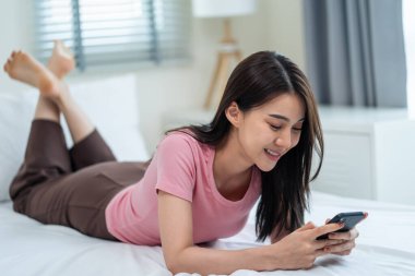 Asyalı çekici kadın sabah evde cep telefonu sohbetini kullanıyor. Mutlu, sıradan, güzel bir kadın yatak odasında uzanıyor. Tatil haftasonunun tadını çıkarın. Evdeki teknolojiyle akıllı telefon iletişimi kurun..