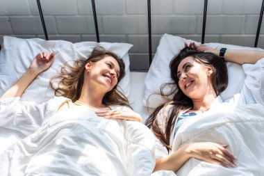 Asian beautiful lesbian couple lying on bed and looking at each other. Attractive romantic girl friend in pajamas spending morning leisure time together in bedroom in house. Homosexual-LGBTQ concept.