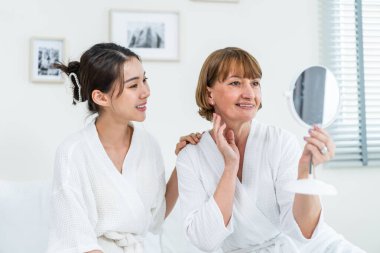 Asyalı kadın annemle yüzüne losyon sürdükten sonra aynaya bakıyor. Çekici genç kız ve bornozlu yaşlı anne yatağa oturur, sağlıklı güzellik ve cilt bakımıyla gurur duyar ve mutlu olurlar..