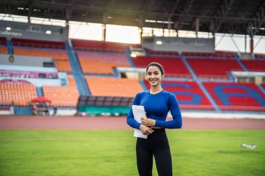 Asyalı aktif spor eğitmeni portresi stadyumda duruyor. Çekici güçlü atlet kız olimpiyat yarışmasında antrenmandan sonra kameraya bakıyor..