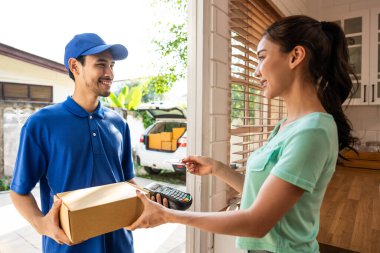 Asyalı genç teslimatçı, evdeki bayan müşteriye paket teslim ediyor. Mavi üniformalı çekici bir postacı çalışıyor, kadına bir kutu teslim ediyor ve kapının önünde kredi kartı ödemesini alıyor.