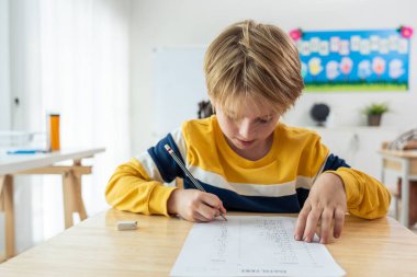 İlkokulda sınav yapan beyaz bir genç çocuk. Sevimli çocuklar masada oturuyorlar, yeni sınıfa bakarken kendilerine güveniyorlar ve anaokulunda öğretmenle birlikte öğreniyorlar.