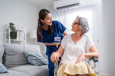 Beyaz, son sınıf öğrencisi kadın, genç Asyalı hemşireyle birlikte tekerlekli sandalyede oturuyor. Çekici kadın bakıcı doktor ziyareti, teşhis koymak ve evdeki oturma odasındaki yaşlı hastayı muayene etmek.