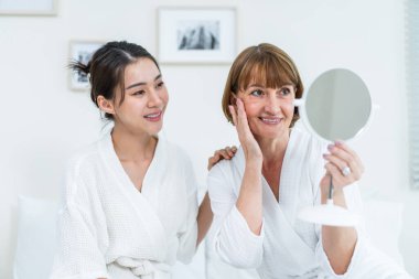 Asyalı kadın annemle yüzüne losyon sürdükten sonra aynaya bakıyor. Çekici genç kız ve bornozlu yaşlı anne yatağa oturur, sağlıklı güzellik ve cilt bakımıyla gurur duyar ve mutlu olurlar..