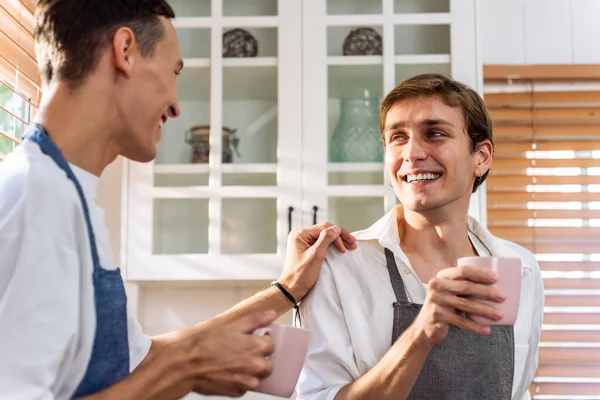 Kafkasyalı genç lgbtq eşcinsel ailesi sabahları bir fincan kahve içer. Çekici, romantik erkek çift önlük giyer. Mutfakta birlikte yemek pişirmek için mutlu ve neşeli olurlar..