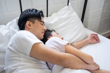 Asian attractive young couple lying down on bed to sleep in bedroom. Beautiful new marriage man and woman in pajamas feel happy and relax on comfortable pillow in the early morning together in house.