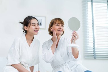 Asyalı kadın annemle yüzüne losyon sürdükten sonra aynaya bakıyor. Çekici genç kız ve bornozlu yaşlı anne yatağa oturur, sağlıklı güzellik ve cilt bakımıyla gurur duyar ve mutlu olurlar..