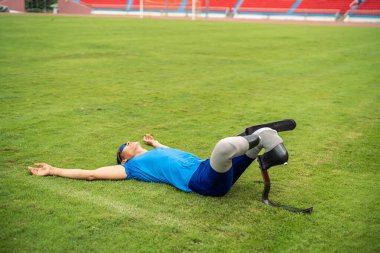 Stadyumda bacak esneten protez bıçaklı Asyalı bir sporcu. Çekici ampüte sporcu koşucusu yerde oturuyor, Engelli Olimpiyatları koşusu antrenmanından sonra mola veriyor..