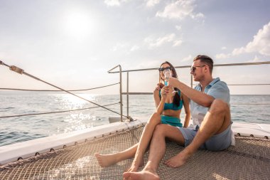 Genç romantik çift yat gezisi sırasında güzel manzaraya bakıyor. Çekici bir genç adam ve kadın birlikte takılıp yıl dönümü balayı gezisini kutlarken katamaran teknesi yaz günbatımında yelken açıyor..
