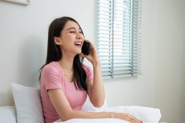 Asyalı çekici kadın sabah evde cep telefonuyla konuşuyor. Mutlu gündelik güzel kadın yatak odasında oturuyor, tatilin tadını çıkarıyor, akıllı telefon kullanarak evdeki teknolojiyle iletişim kuruyor..