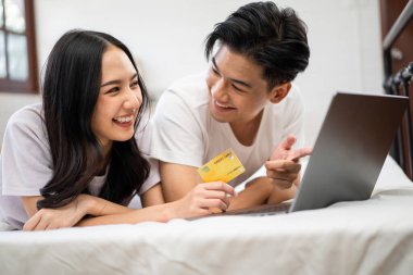 Asyalı çekici kadın sabahları kocasıyla yatakta dizüstü bilgisayar kullanıyor. Birlikte uzanan yeni evli çift yatak odasında uyandıktan sonra internet üzerinden alışveriş yaparken mutlu ve rahatlamış hissediyorlar.