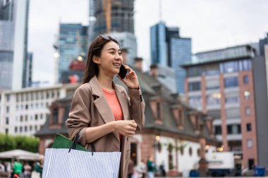 Mağazada yürürken Asyalı güzel kadınlar telefonda konuşuyor. Alışveriş çantası tutan ve mutlulukla yürüyen çekici bayan gezgin, Frankfurt 'taki alışveriş merkezinden satın almanın keyfini çıkarın.
