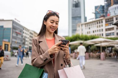 Asyalı güzel kadınlar mağazada yürürken akıllı telefon kullanıyorlar. Çekici genç bir bayan alışveriş çantası tutuyor ve sonra mutlulukla yürüyor. Alışveriş merkezinde birlikte alışveriş yapmanın tadını çıkarıyorlar..