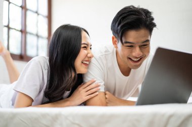 Asyalı çekici kadın sabahları kocasıyla yatakta dizüstü bilgisayar kullanıyor. Yeni evli çiftimiz yatak odasında uyandıktan sonra bilgisayardan film izlerken mutlu ve rahatlamış hissediyorlar.