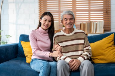 Evde kızıyla birlikte kanepede oturan Asyalı yaşlı babanın portresi. Çekici güzel torun mutlu ve rahat hissediyor, olgun büyükbabayı ziyaret ediyor ve evde kameraya bakıyor..
