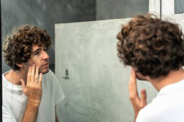 Beyaz, genç bir adam yüzünü köpük ve suyla yıkıyor. Çekici erkek, sağlıklı güzellik tedavileri ve cilt bakımı için yüzünü yıkıyor sonra aynaya bakıyor ve banyoda gözlük takıyor..