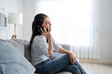 Asyalı güzel kadın kanepede oturur ve cep telefonuyla konuşur. Mutlu çekici genç kız evde vakit geçirir, rahatlar, iletişim kurar ve oturma odasında akıllı telefondan tartışır..
