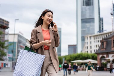 Mağazada yürürken Asyalı güzel kadınlar telefonda konuşuyor. Alışveriş çantası tutan ve mutlulukla yürüyen çekici bayan gezgin, Frankfurt 'taki alışveriş merkezinden satın almanın keyfini çıkarın.