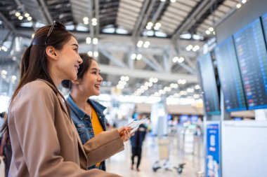 Asyalı genç bayan yolcular havaalanındaki uçuş kartlarını kontrol ediyorlar. Çekici güzel bayan turist arkadaşları tatil gezisi için uçakla yurtdışına gitmekten mutlu ve heyecanlı hissediyorlar..