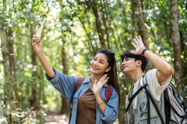 Asyalı genç bir çift ormanda selfie çekiyor. Çekici erkek ve kadın arkadaş gezgini telefon kaydı video bloğu kullanıyor, tatil boyunca doğa tahtasında mutlulukla yürüyorlar.