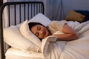 Asian beautiful woman lying down on bed in bedroom, dreaming at night. Attractive young female in pajamas sleeping alone on comfortable pillow and cozy blanket in dark room for health care at home.