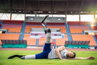 Stadyumda bacak esneten protez bıçaklı Asyalı bir sporcu. Çekici ampüte sporcu koşucusu yerde oturuyor, Engelli Olimpiyatları koşusu antrenmanından sonra mola veriyor.