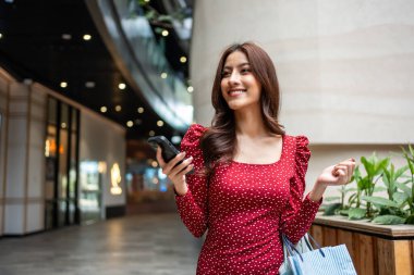 Asyalı güzel kadınlar mağazada yürürken akıllı telefon kullanıyorlar. Çekici genç bir bayan alışveriş çantası tutuyor ve sonra mutlulukla yürüyor. Alışveriş merkezinde birlikte alışveriş yapmanın tadını çıkarıyorlar..