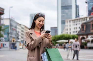 Asyalı güzel kadınlar mağazada yürürken akıllı telefon kullanıyorlar. Çekici genç bir bayan alışveriş çantası tutuyor ve sonra mutlulukla yürüyor. Alışveriş merkezinde birlikte alışveriş yapmanın tadını çıkarıyorlar..